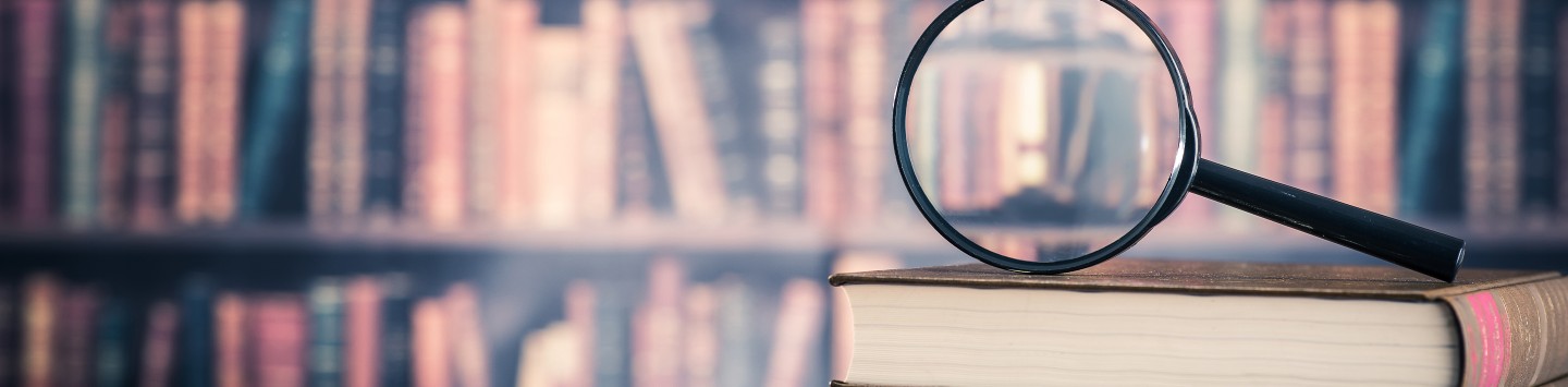 Cropped 2880x1100 of library with books in background and magnifying glass on top of books in the foreground